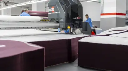 Automated blade cutting intricate patterns into deep stacks of dark fabric covered with cutting paper.