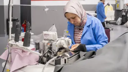 Female worker in a blue coat sewing bright pink garments at her machine.