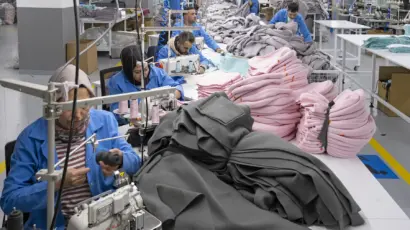 Female machinist in a blue factory uniform expertly stitching dark fabric.